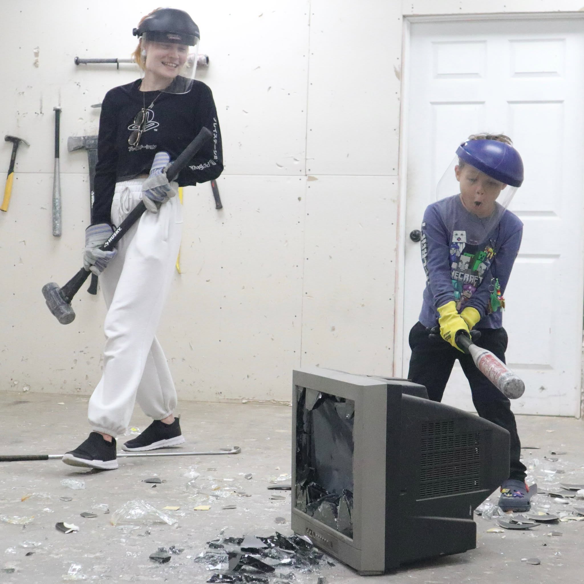 A parent and child in safety gear breaking an old TV together in a private smash session
